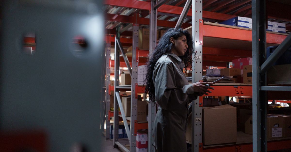An adult woman in uniform checking inventory in a warehouse aisle.