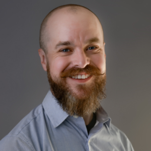Shaun Brown, Senior ERP Implementation Manager at GVO, smiling with a beard, wearing a light blue shirt, representing expertise in AI, compliance, and ERP systems for the webinar