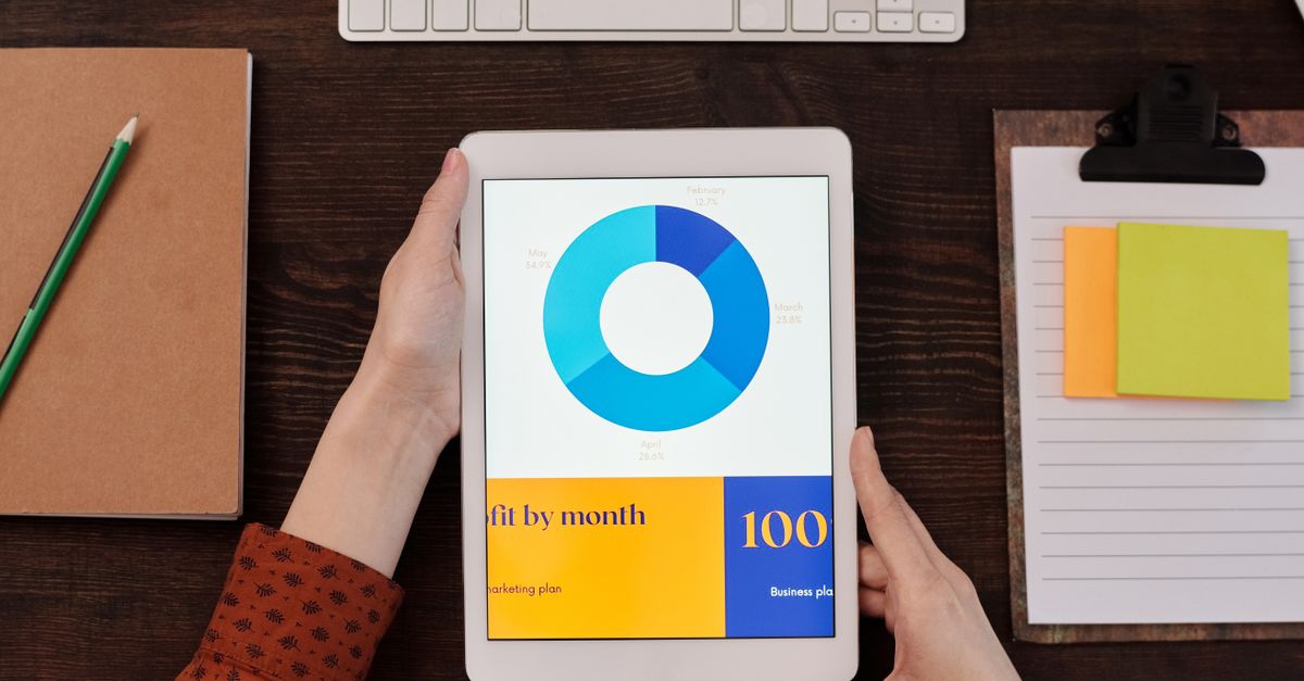 Tablet showing pie chart on desk with hands, keyboard, and notepad in office setting.