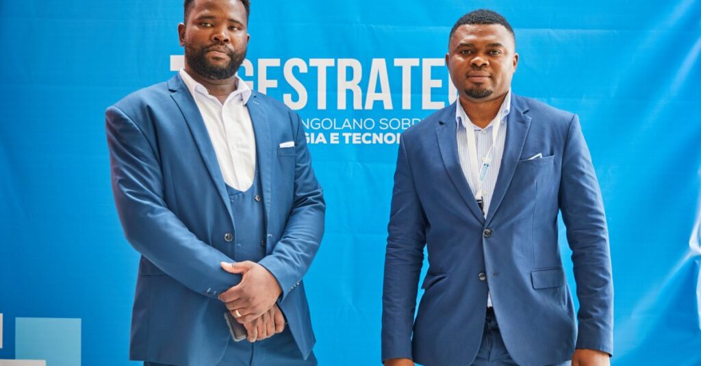 Two men in blue suits pose at a professional conference against a branded backdrop.
