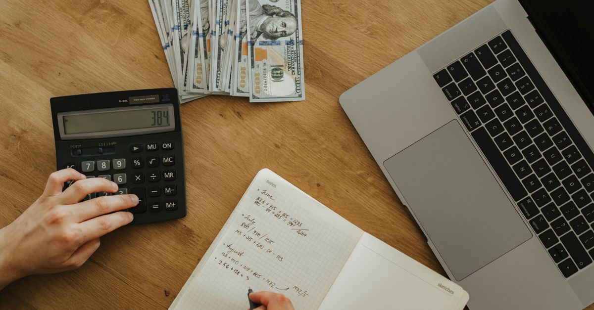 Hands using calculator and notebook, planning finances, with cash and laptop on wooden table.