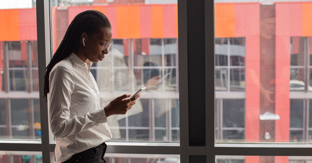 African American woman focused on her smartphone by a large office window, urban background.