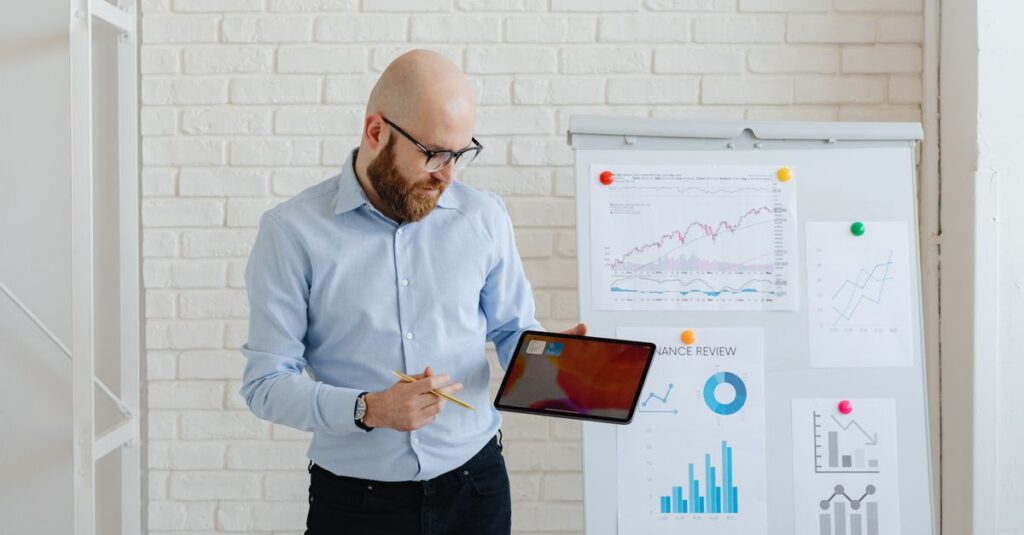 Bald man presenting finance data with tablet and graphs on whiteboard.