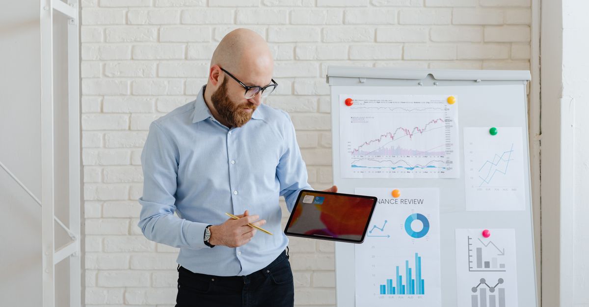 Bald man presenting finance data with tablet and graphs on whiteboard.