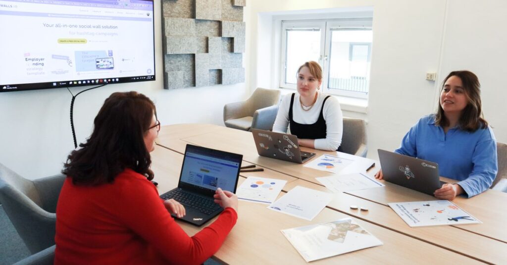 Three women collaborating at a conference table in a modern office with laptops and presentation screen.