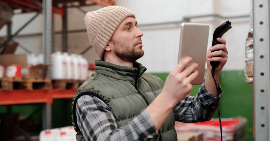Man in beanie and vest using a scanner in a warehouse for inventory control.