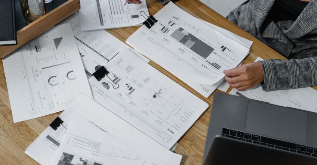 Woman analyzing financial data and reports on a wooden desk with a laptop, showcasing analytics in a workspace.