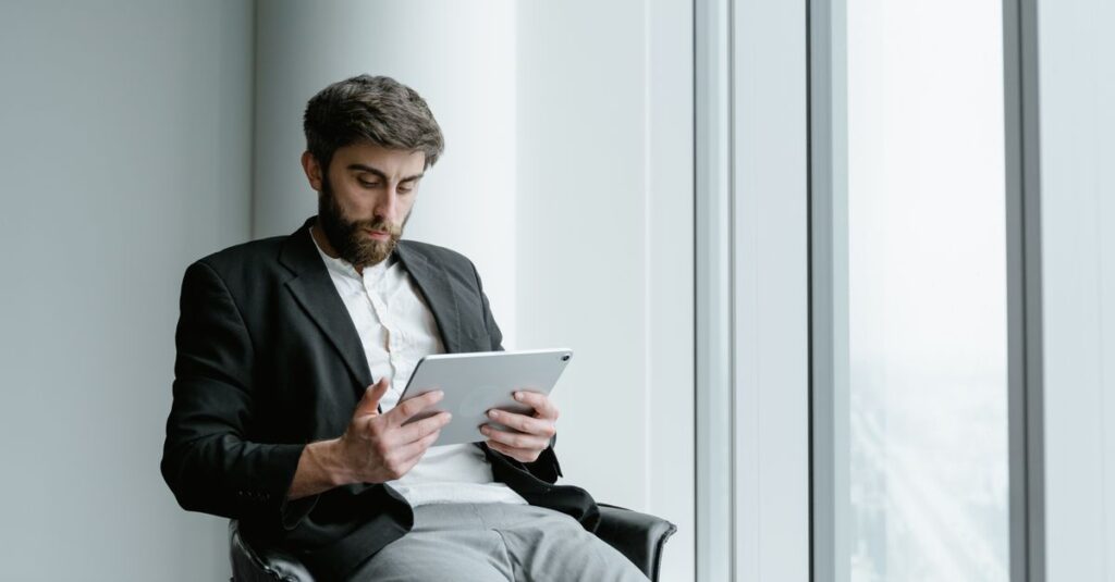 A man in formal attire working on a tablet while seated by a large office window.
