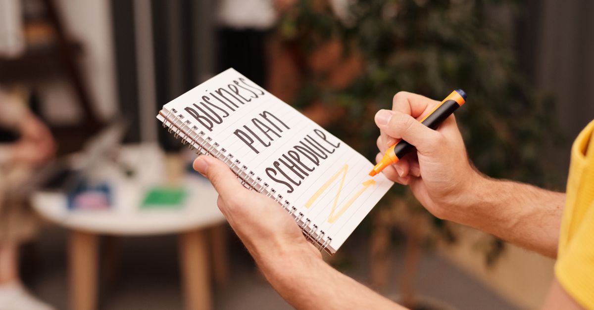 Close-up of a hand writing in a notebook with 'Business Plan Schedule' indoors.