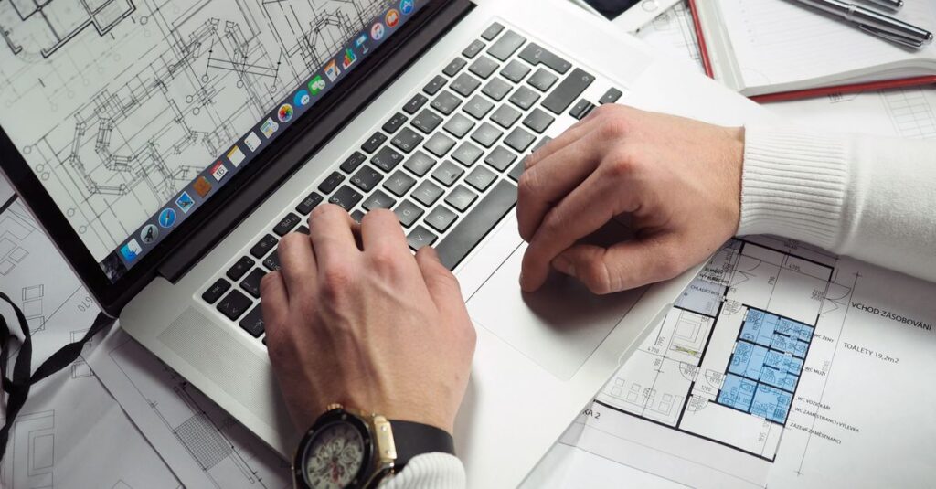 Architect reviewing detailed floor plans and schematics on a laptop with a smartphone nearby.