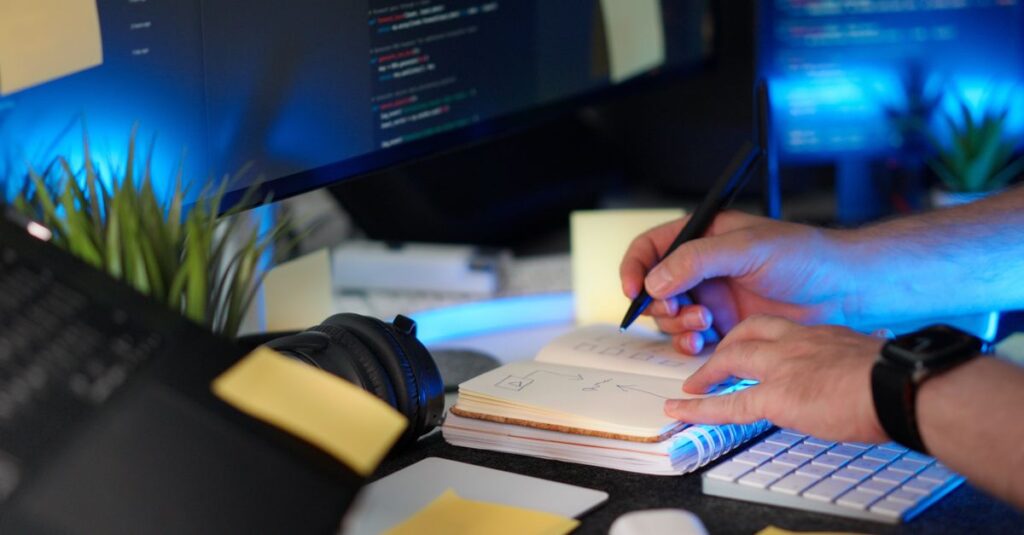 A person planning a software project with handwritten notes and code on a monitor in a modern workspace.