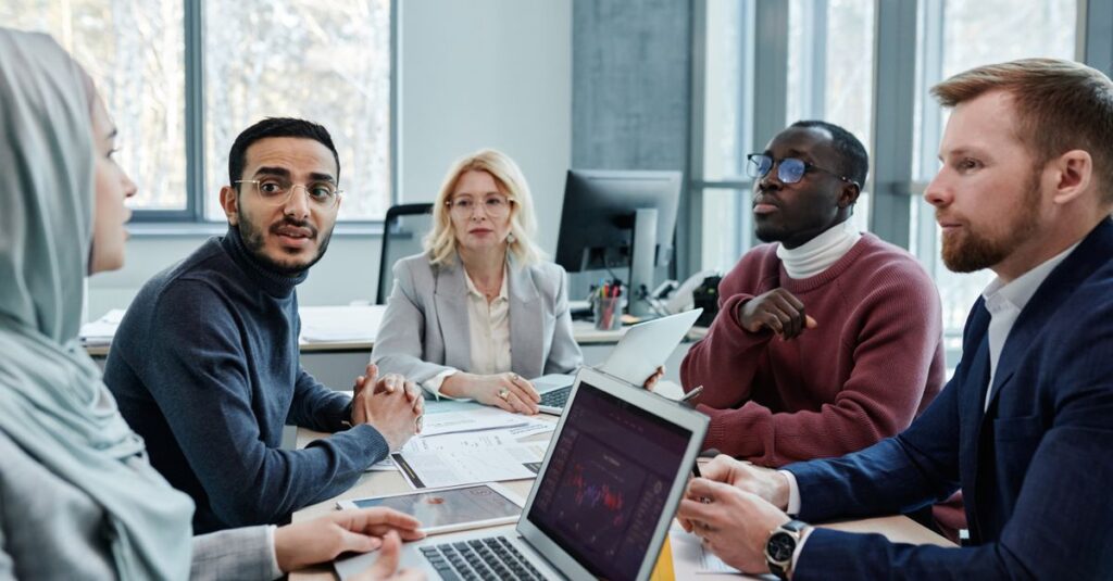 Diverse group of professionals engaged in a conference room discussion with laptops and documents.