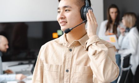 Asian man in beige shirt smiling with headset in modern office setting.