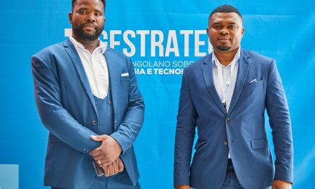 Two men in blue suits pose at a professional conference against a branded backdrop.