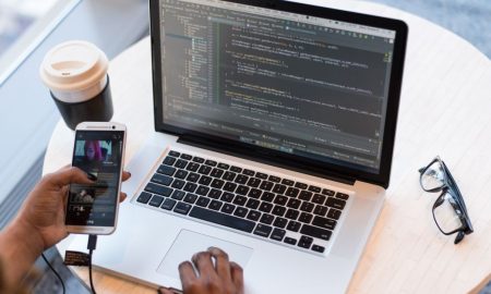 A programmer manages code on a laptop while multitasking with a smartphone, illustrating modern tech workspace.