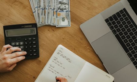 Hands using calculator and notebook, planning finances, with cash and laptop on wooden table.