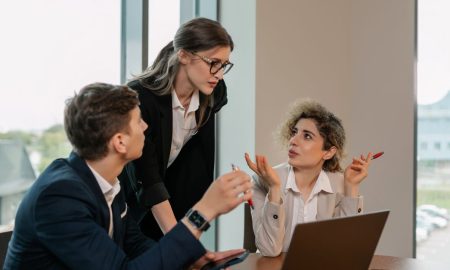 Three professionals in a business meeting, discussing work around a laptop in a modern office.