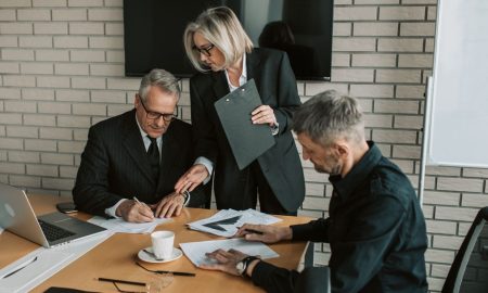 Three mature professionals in a business meeting discussing and signing documents in an office setting.