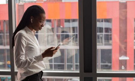 African American woman focused on her smartphone by a large office window, urban background.