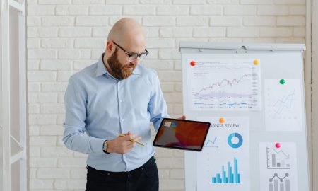 Bald man presenting finance data with tablet and graphs on whiteboard.