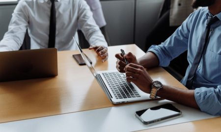 Professionals in a corporate meeting room engaged in discussion with laptops and smartphones.