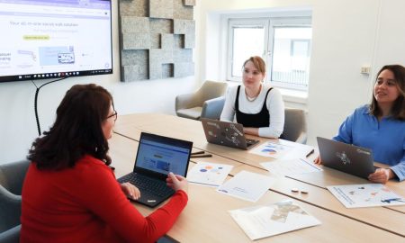 Three women collaborating at a conference table in a modern office with laptops and presentation screen.