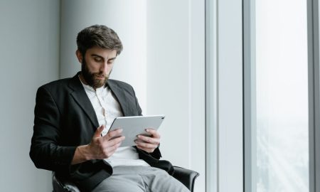 A man in formal attire working on a tablet while seated by a large office window.