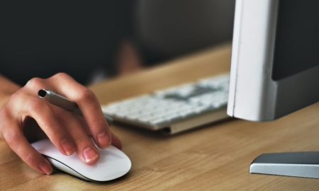 A hand using a wireless mouse at a modern desk setup with a computer and keyboard.