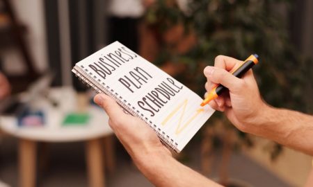 Close-up of a hand writing in a notebook with 'Business Plan Schedule' indoors.